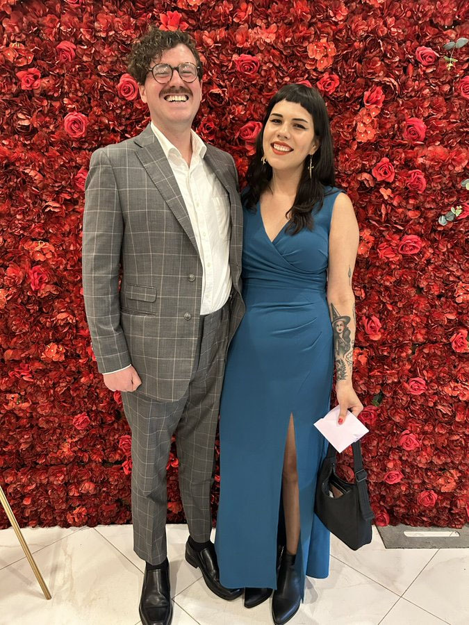 Ryan Carson and his girlfriend pose in formal attire in front of a red rose backdrop at a wedding. 