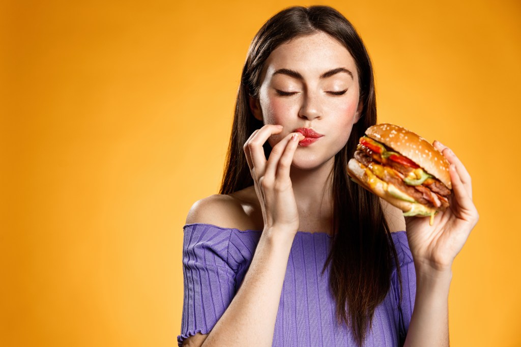 Woman eating cheeseburger with satisfaction. Girl enjoys tasty hamburger takeaway, licking fingers delicious bite of burger, order fastfood delivery while hungry, standing over orange background.