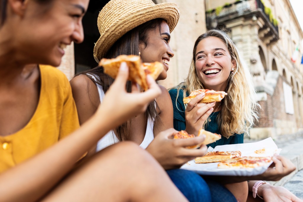 Three young multiracial women laughing while eating a piece of pizza in italian city street - Happy female friends enjoying holidays together in Italy - Friendship, travel and tourism concept