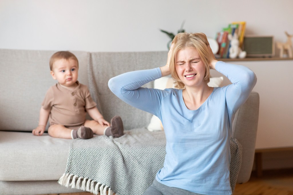 Stressed blonde woman young mother suffering from headache, sitting with baby at home, touching her head and screaming. 
