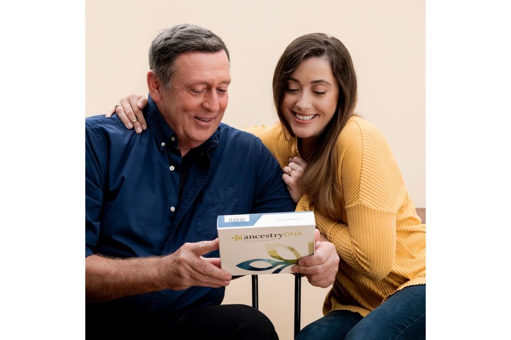 Two people sitting next to each other; one holding an AncestryDNA kit.