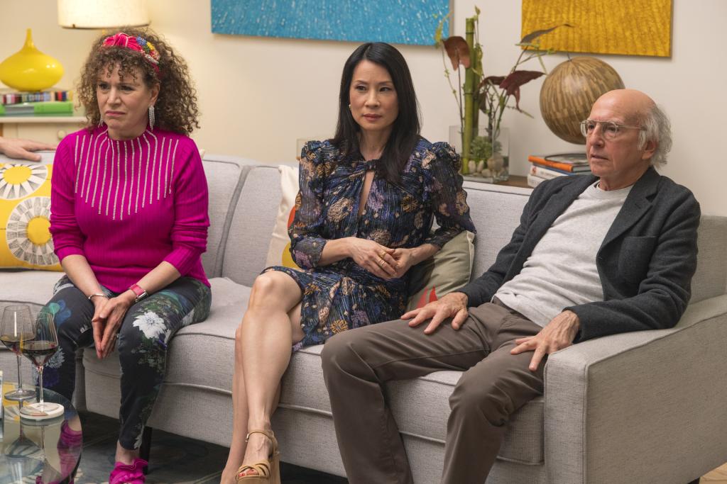 Susie Essman, guest star Lucy Liu and Larry David in a scene from "Curb Your Enthusiasm." They're seated on a couch next to each other. There are two wine glasses on a glass table in front of them. Larry has his arms on his thighs and they all look bemused.