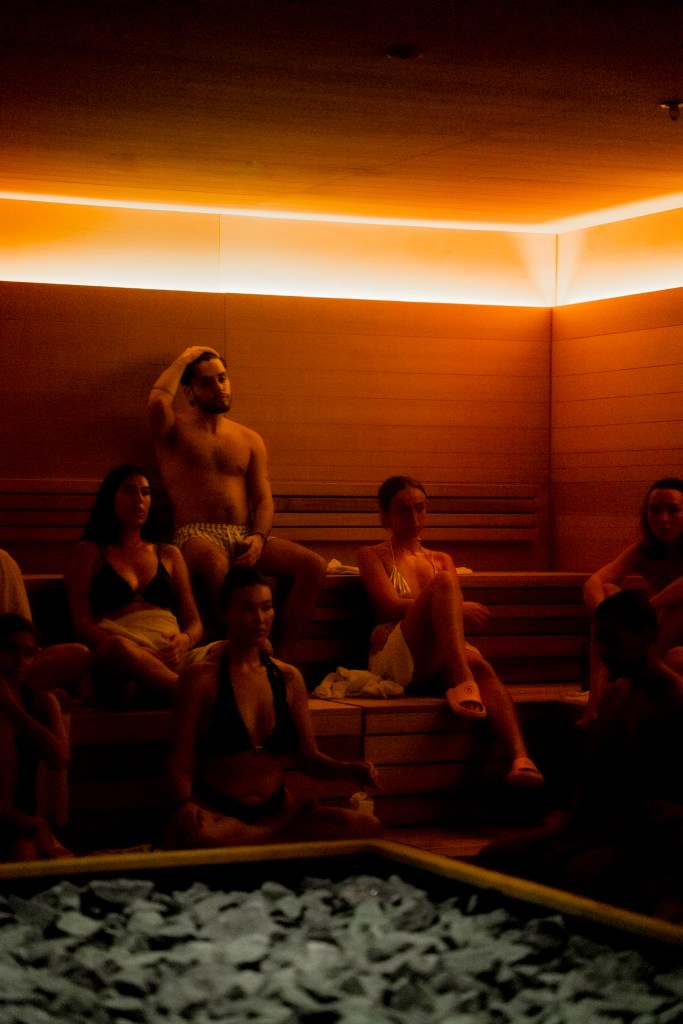 One man and three women, all in bathing suits, sit in the sauna at Bathhouse Flatiron. In the foreground, coals can be seen. Glowing lights are seen at the top of the frame. 