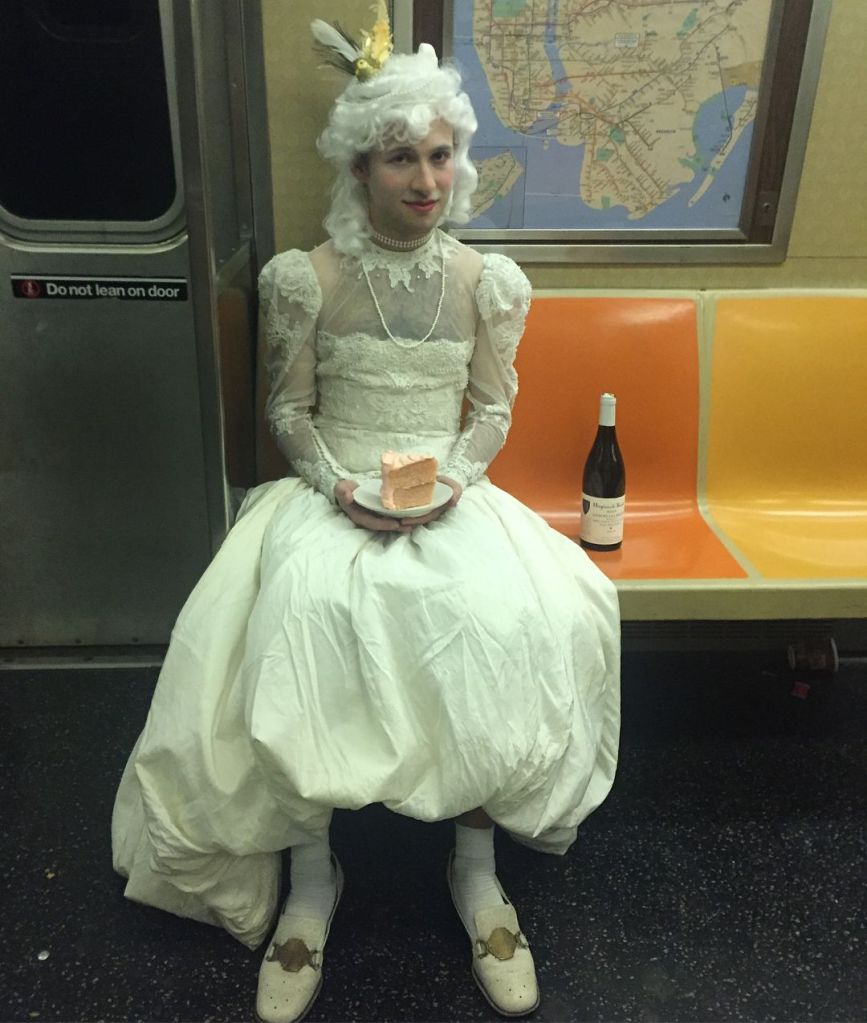 Eli Coplan on the subway with a cake, white wig and bottle of wine.