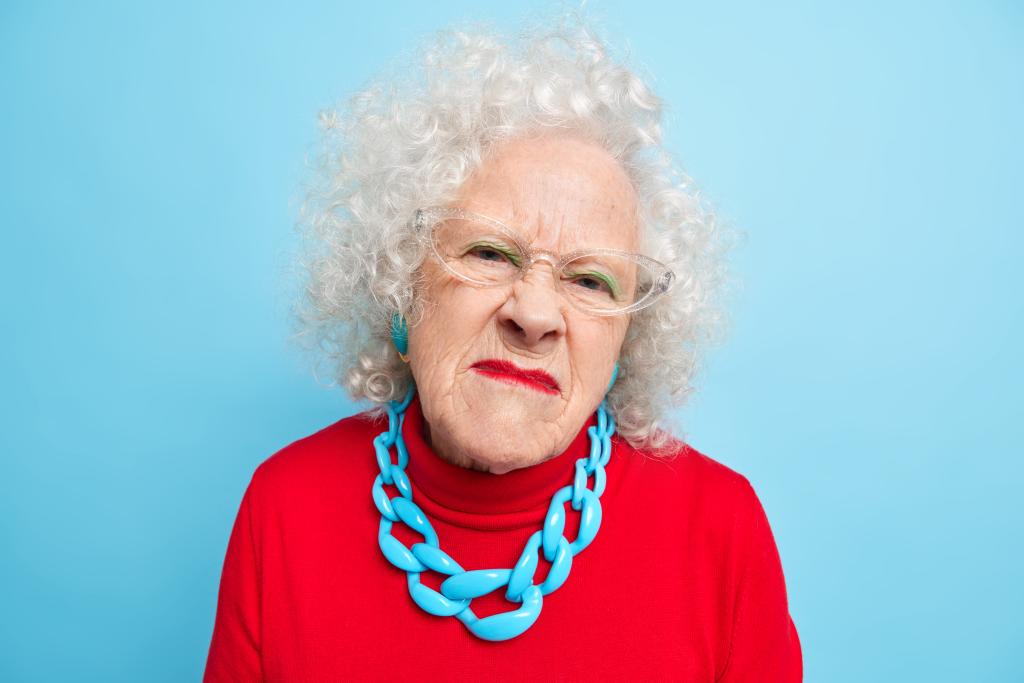 Portrait of displeased senior woman looks with unhappy annoyed face expression at camera dislikes news wears transparent glasses red jumper and necklace isolated over blue background. Studio shot