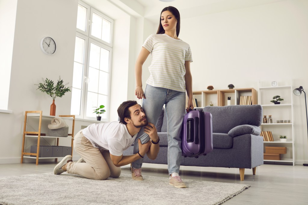 Young couple breaking up. Woman with packed bag leaving home. Funny clingy traitor husband on floor holding wife's leg imploring her to give second chance. Relationship breakup and divorce concept