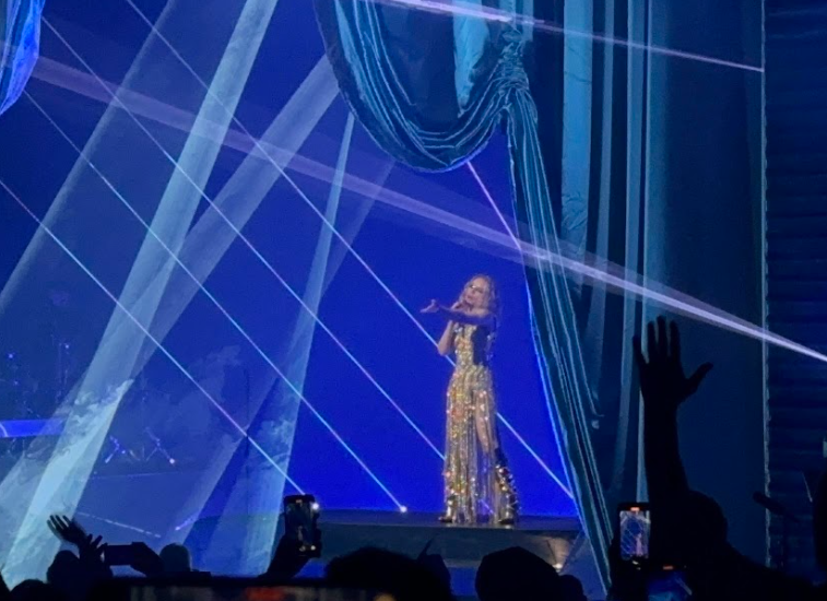 Kylie Minogue addresses the crowd while singing in front of a blue backdrop.