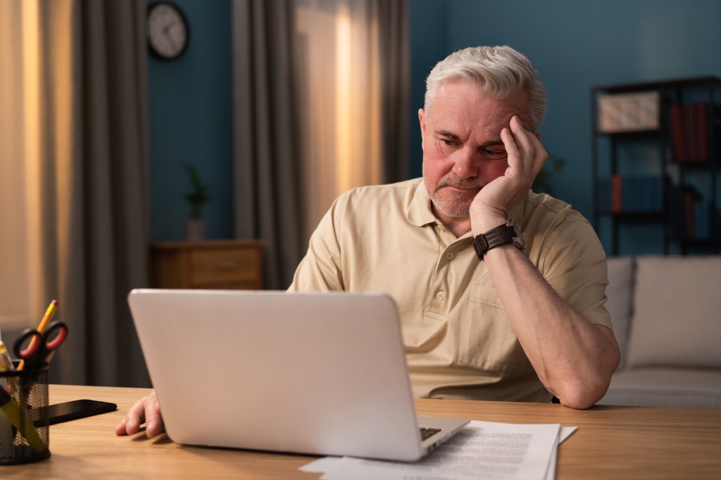 Elderly man looking tired and supporting his head with his hand while sitting in front of a laptop