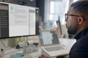 Stock image showing a black man’s face looking into a computer screen in an open plan working office. Type is being added to the screen by an Artificial intelligence, AI, chatbot.