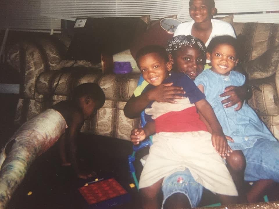 Two young girls sit together on a couch, one in a blue dress. This is a childhood photo of Kennedy Sanders and her best friend Quoyah McPherson.