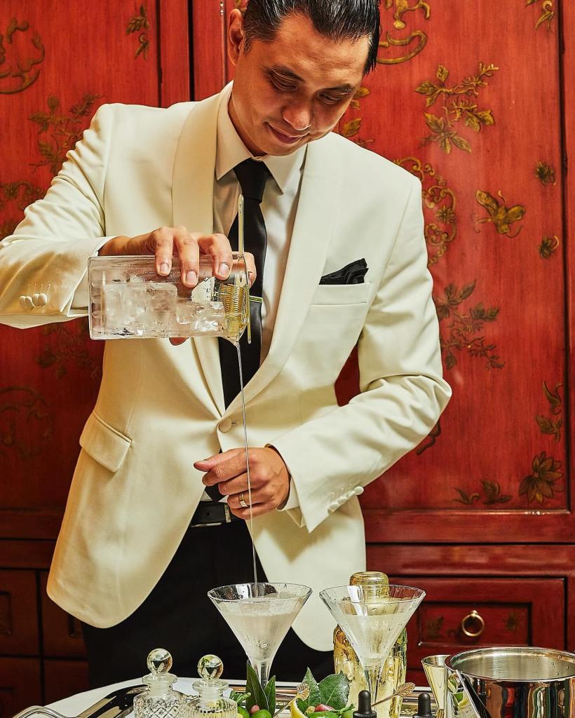 Darryl Chan in a white suit pouring a martini into martini glasses on bar cart.