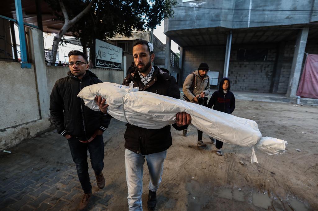 A man carries the body of an individual killed in an attack, wrapped in a white shroud, with another man walking next to him and a third, along with a boy, in the background.