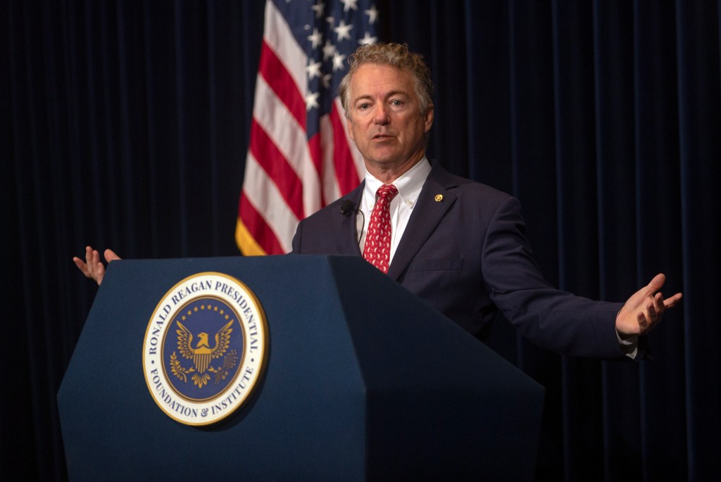 Senator Rand Paul speaking at a podium with a flag backdrop.