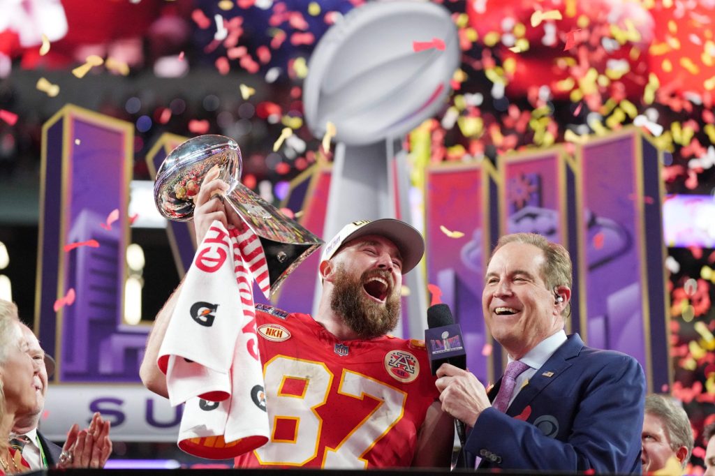 Travis Kelce holding Super Bowl trophy while being interviewed by Jim Nantz at Allegiant Stadium after KC Chiefs win over SF 49ers.