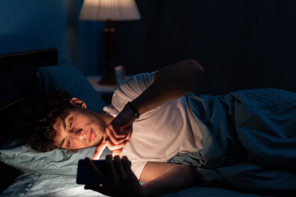 Portrait of young sleepy man lying in bed using smartphone at late night, no sleep. Insomnia, sleep disorder concept. Dependency on a cellphone. Loneliness crisis. Copy space