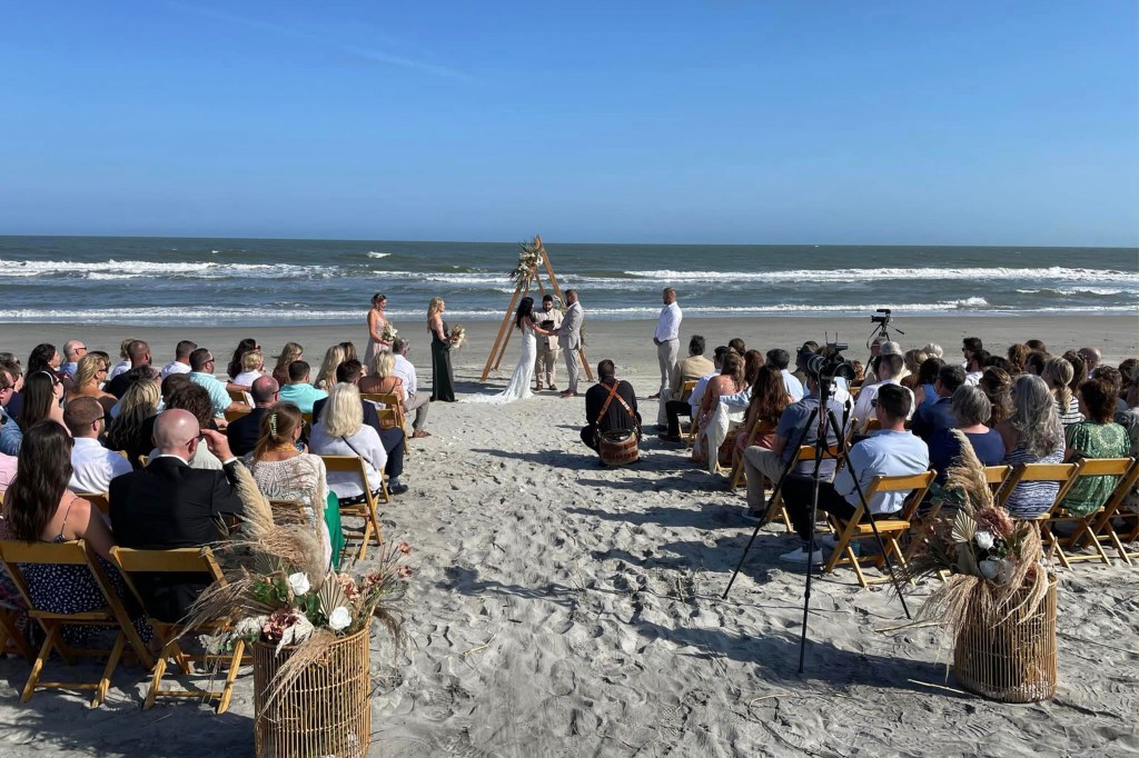 Samantha Miller and Aric Hutchinson hold hands while saying their vows at their beachside wedding as dozens of guests watch on.