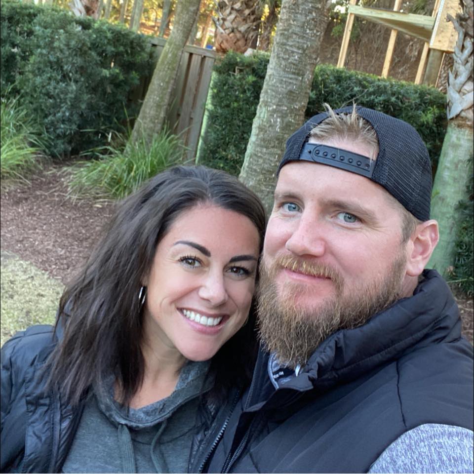 Samantha Miller and Aric Hutchinson are dressed casually and lean in together and smile while standing outside with trees behind them