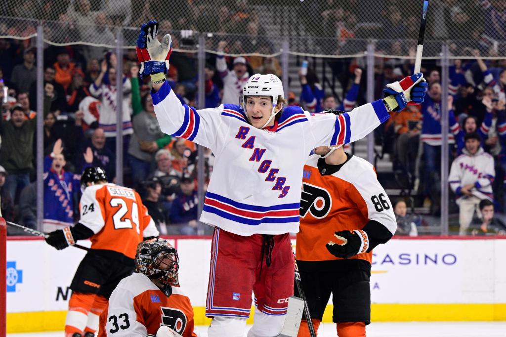 Hockey player in white jersey with raised arms celebrates after scoring past goalie in orange jersey on ice rink during a game.