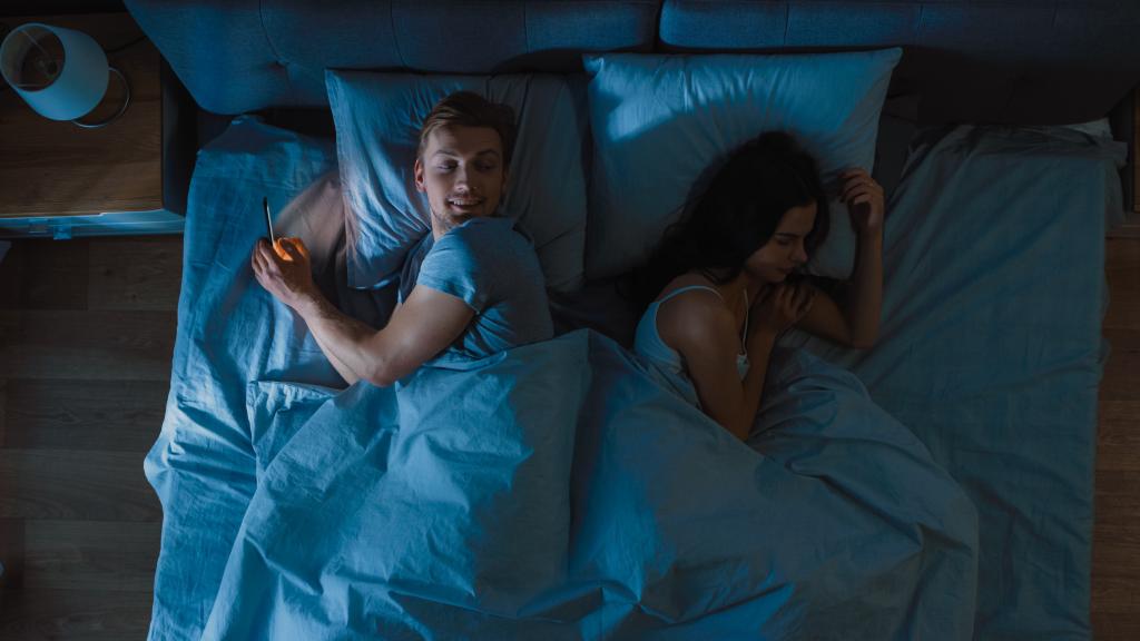 A man using smartphone while girlfriend sleeps in bed beside him.