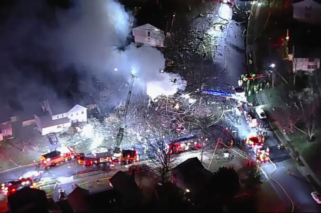 At least a half-dozen emergency vehicles line the road as smoke rises from a property where a home exploded. Other homes nearby appear undamaged.