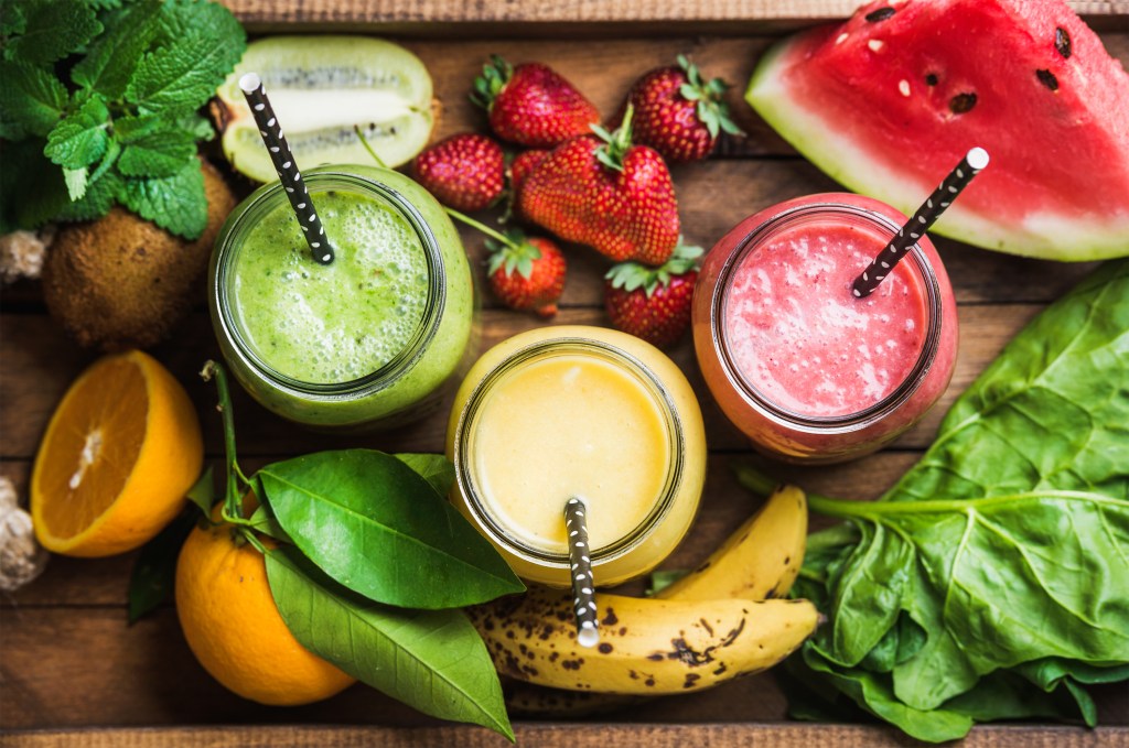 Freshly blended fruit smoothies of various colors and tastes in glass jars in rustic wooden tray. Yellow, red, green. Top view, selective focus