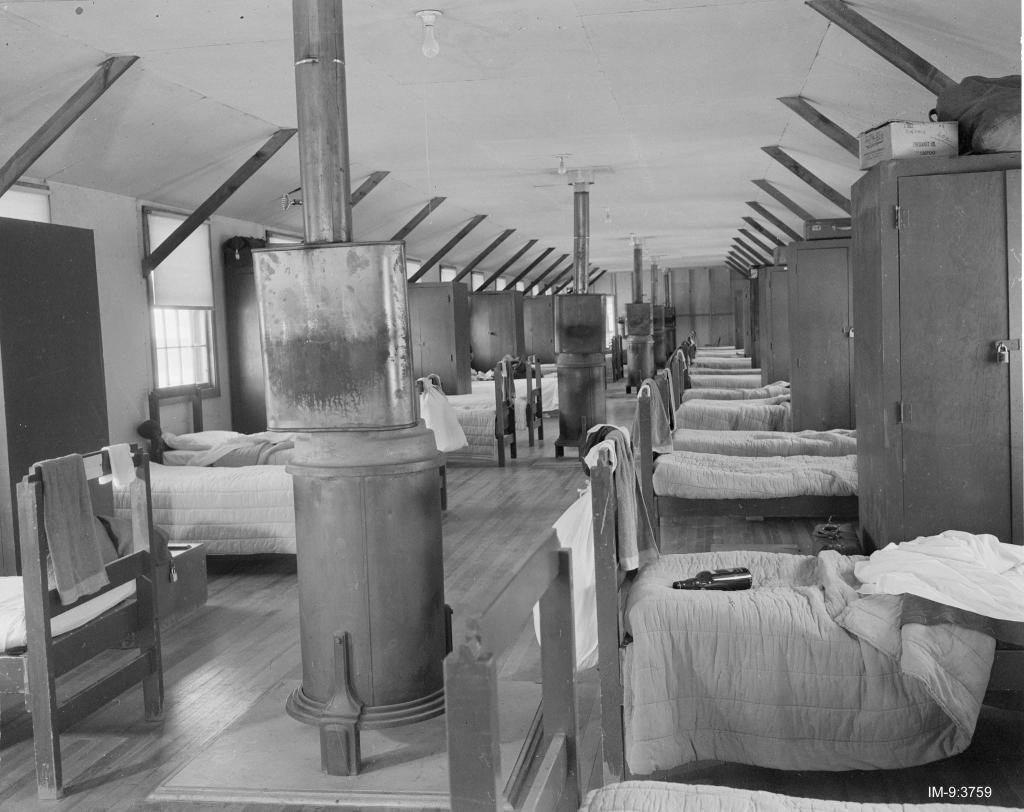 The interior of an army barracks with rows of beds, lockers, and wood-fire stoves in the center aisle.