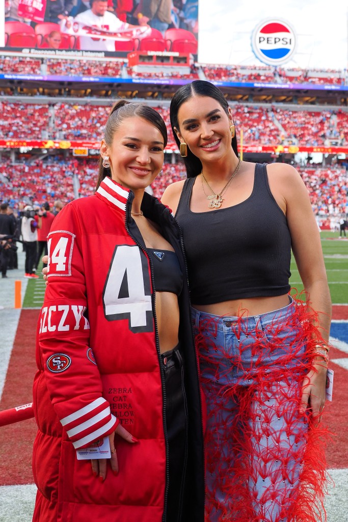 Kristin Juszczyk and Claire Kittle standing together in a stadium, Kristin wears custom made jacket to show support for her husband.