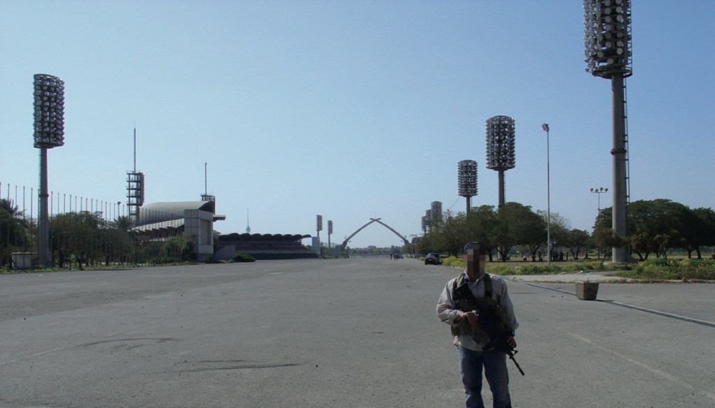 Adam Gamal, with blurred face, stands in front of Saddam Husseim's "Victory Arch" while holding a large gun. 