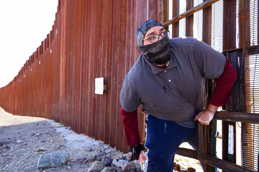 A man wearing a face mask and cap climbs through a gap in the border wall