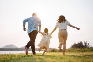 Happy family running on grass in the park during sunset with river in the background, parents holding child's hand.