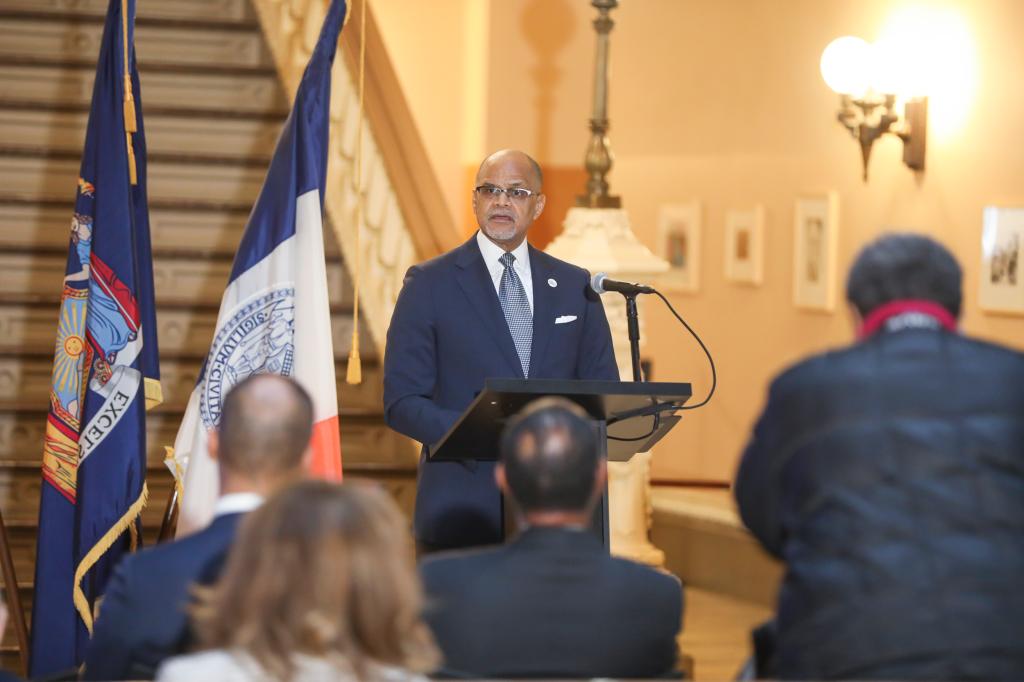 A man speaking at a podium with people seated in front of him, holding a microphone.