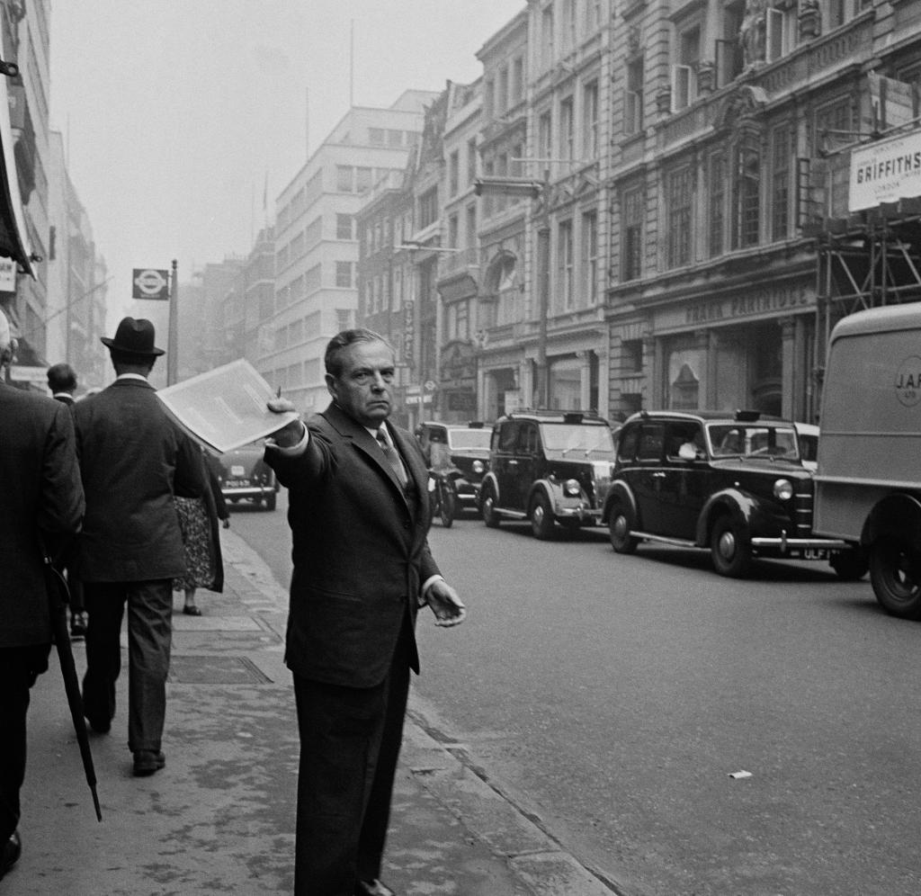 Black-and-white photo of Harry Winston standing on a street in London and holding up papers.