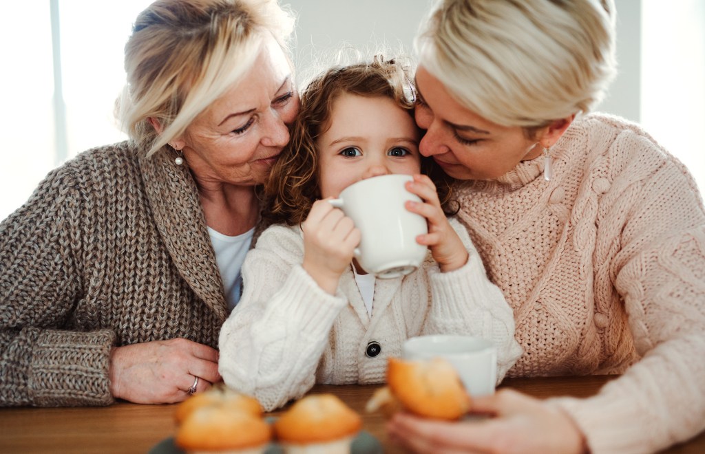 A happy small girl with her mother and grandmother sitting at a table at home, drinking from a mug.