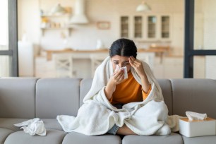 Ill indian woman blowing her nose with tissue on a couch, wrapped in a blanket, showing signs of cold and flu.