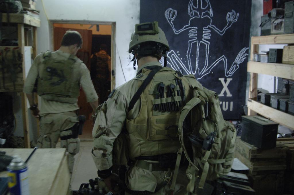 Two men in military gear walking out a door, seen from behind. 