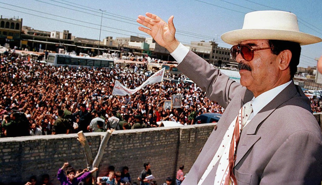 Saddam Hussein wearing a suit and hat, waving to crowds in Baghdad.