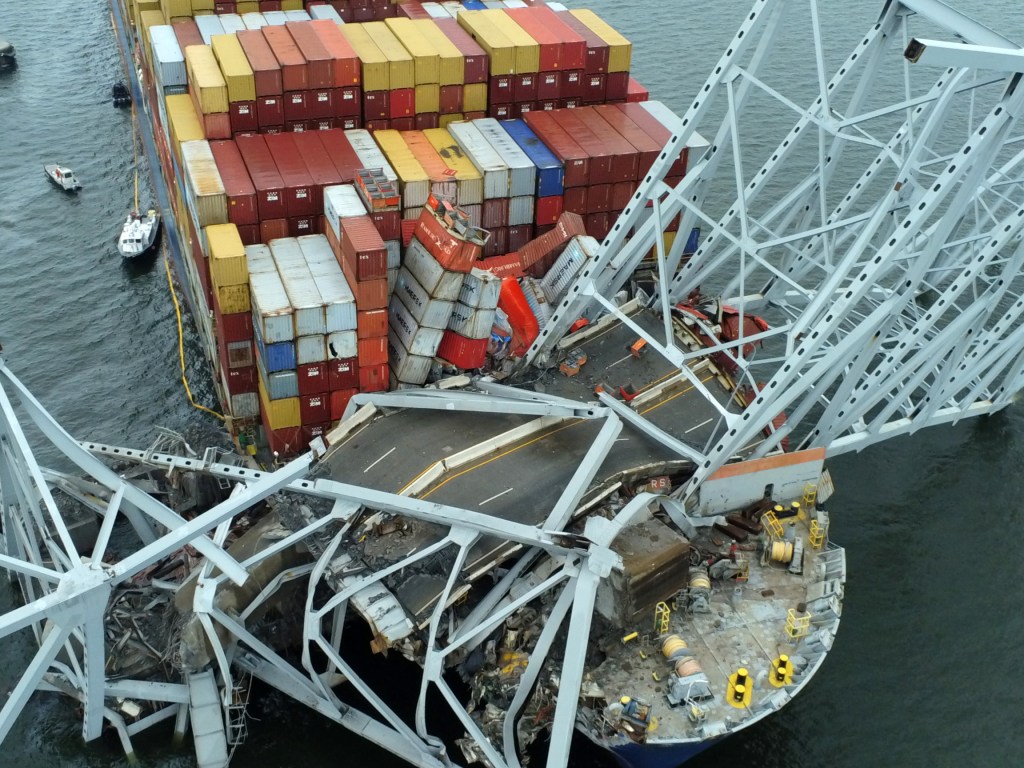 A view from above of  damaged girders and roadway from the collapsed Francis Scott Key Bridge in Baltimore atop the M.V. Dali, the ship that crashed into the bridge and caused its collapse. Hundreds of red, yellow, blue and white shipping containers can be seen on the deck of the ship as well.