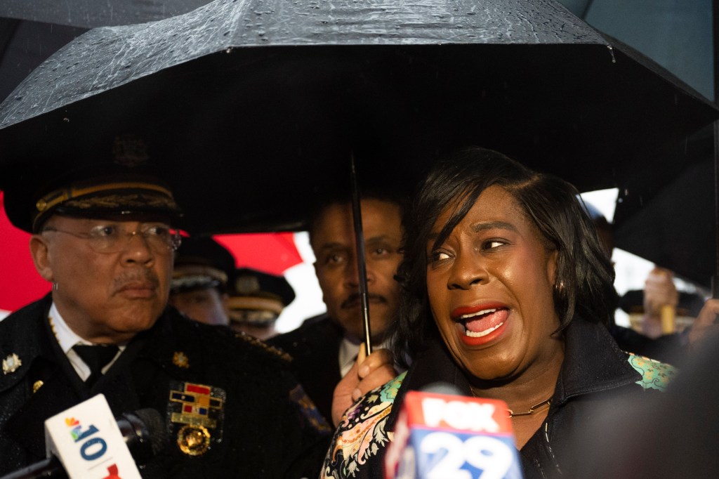 Philadelphia Mayor Cherelle Parker speaks to the media in front of a building with an umbrella while holding a microphone.