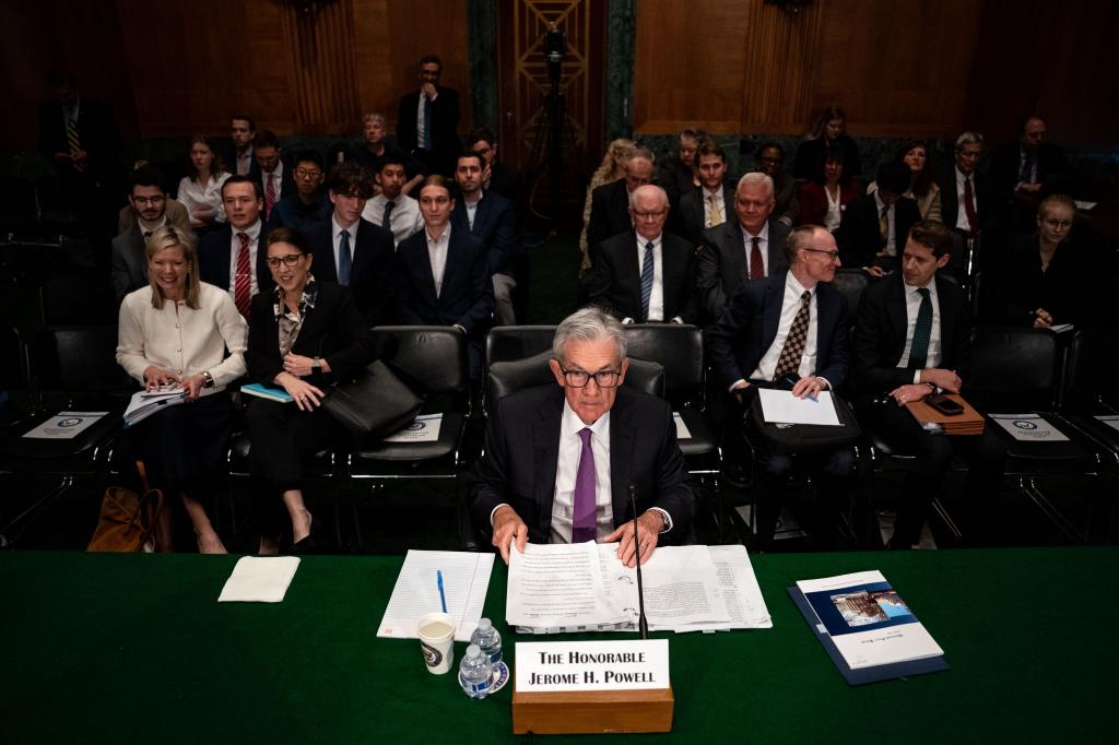 Jerome Powell testifies during Senate Banking hearing, surrounded by people; uncertain economic outlook.