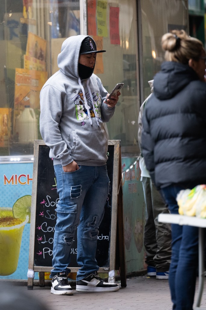 A man who is selling fake ID cards standing on the street looking at a his phone. 