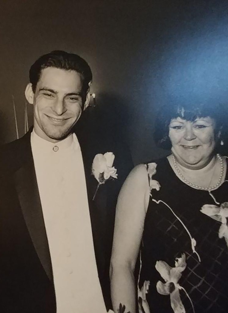 Frank Meek and his mother, Thomasine (right). Frank is wearing a tuxedo with a rose buttonhole arrangement and his mother is wearing pearls and a sleeveless dark dress with a large flower print.