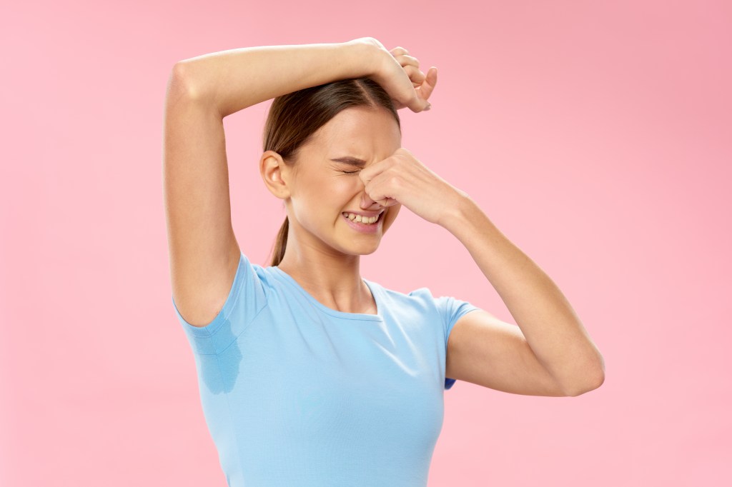 A woman with sweat stain in armpit, holding nose making disgusted face with arm up, against a pink background