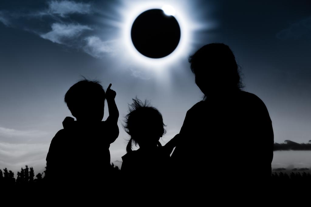 Natural phenomenon. Silhouette back view of mother and child sitting and relaxing together. Boy pointing to solar eclipse on dark sky background. 