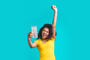 Excited attractive dark skinned girl in bright yellow t-shirt holding lottery ticket in hand and making winner gesture on blue background screaming and laughing celebrating victory.