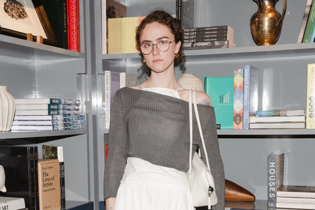 Ella Emhoff, in glasses wearing a grey sweater over a white dress with a white purse on her shoulder, at La Mercerie