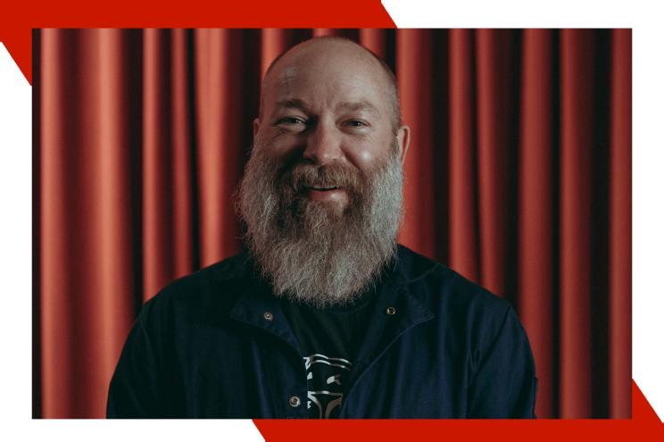 Bearded comedian Kyle Kinane smiles in front of a red curtain.