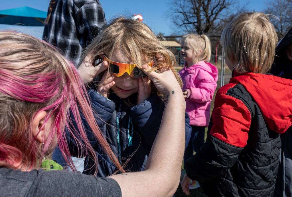 A young woman helps a child put eclipse glasses on their face.