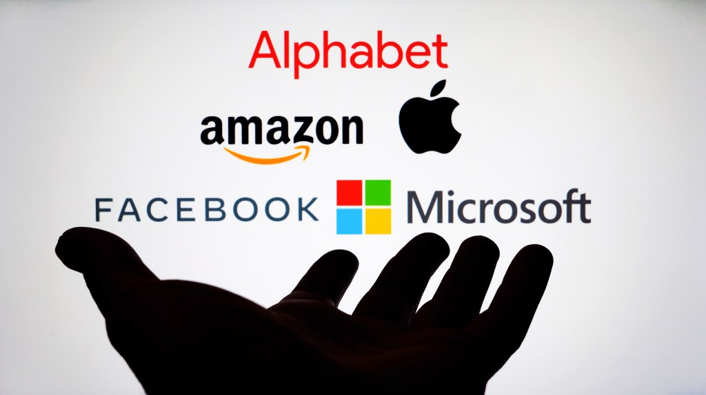 Logos of Alphabet, Amazon, Apple, Facebook, and Microsoft on a screen with a hand reaching out. Addressing digital tax and antitrust. 