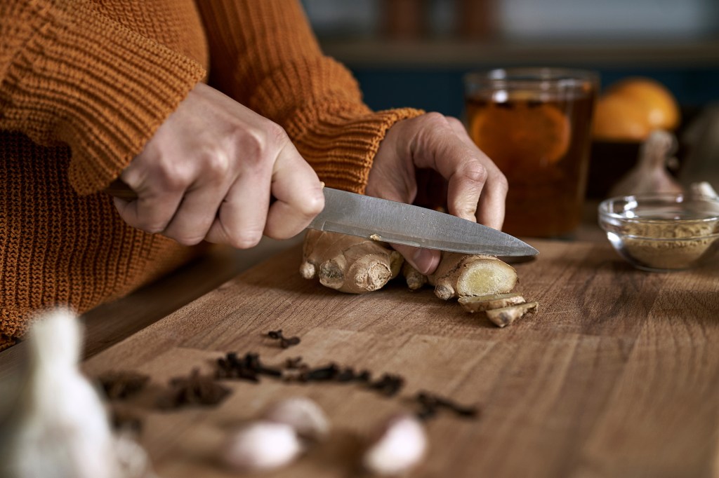 A caucasian woman cuts ginger on a wooden board in the kitchen, captioned as a potential cure to a stuffy nose.