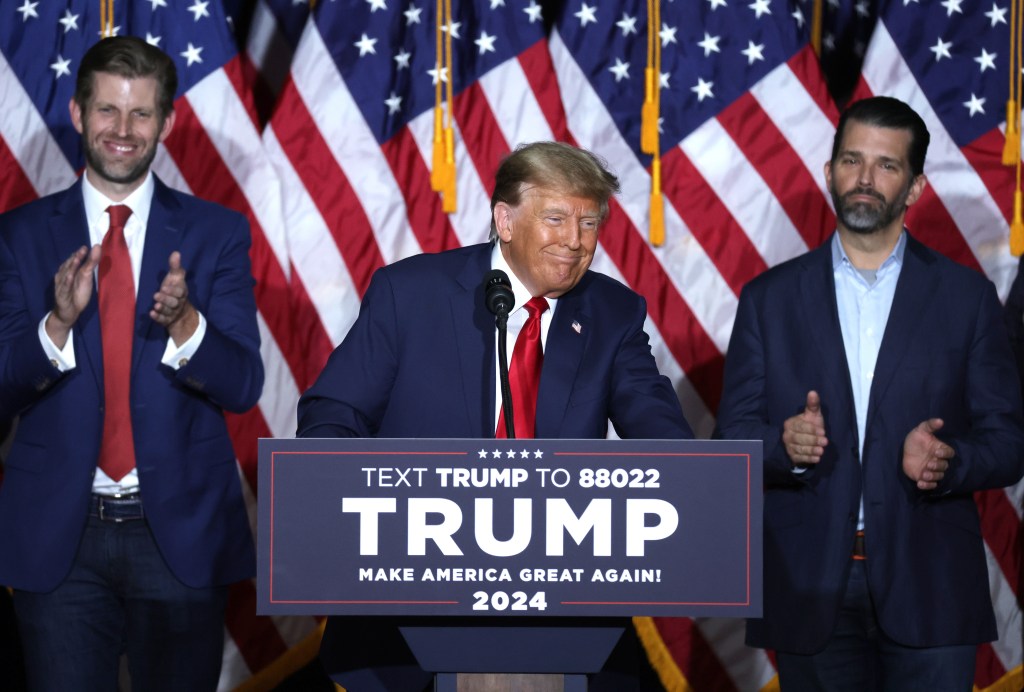 Former President Donald Trump speaking at a podium during an Iowa caucus night event, with sons Donald Trump Jr. and Eric Trump in the background.
